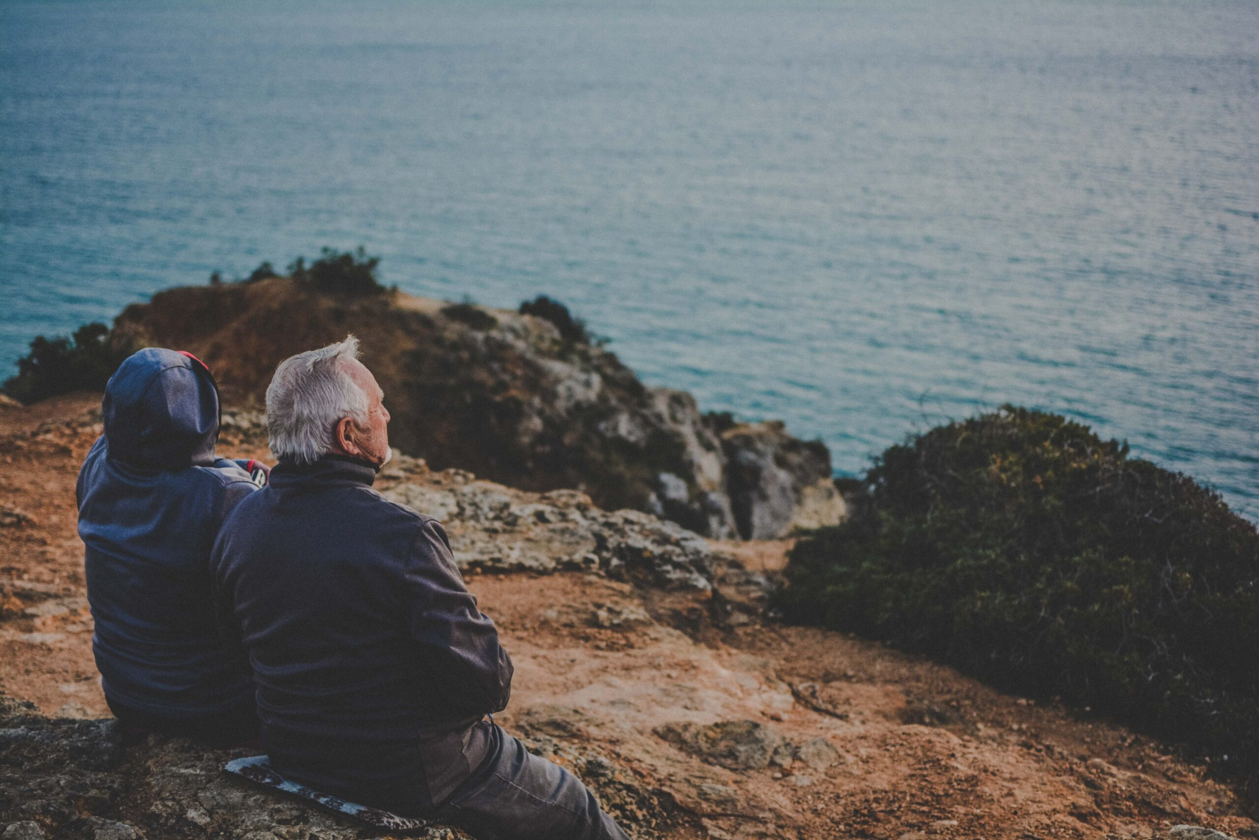 Two people sitting on a rocky cliff overlooking the ocean.