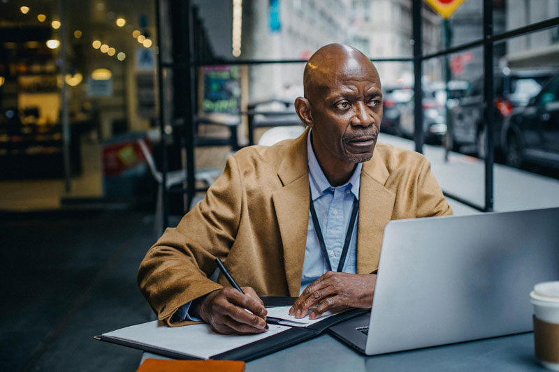 Man working on a laptop and taking notes at an outdoor cafe.