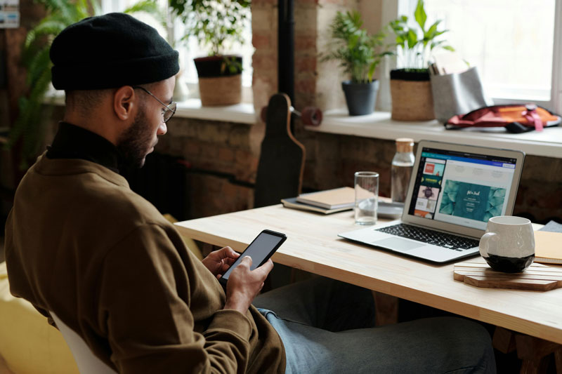 Man sitting at a desk, using a smartphone with a laptop open in front of him.