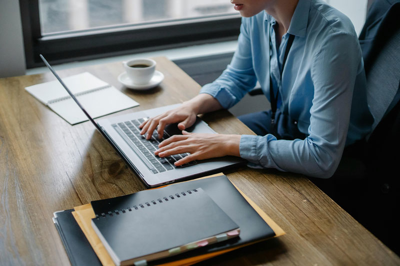 Woman typing on a laptop at a desk with notebooks and a cup of coffee.