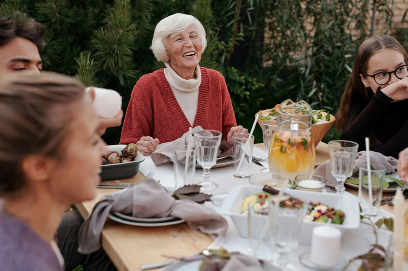 Elderly woman laughing while seated at an outdoor dining table with family and friends.