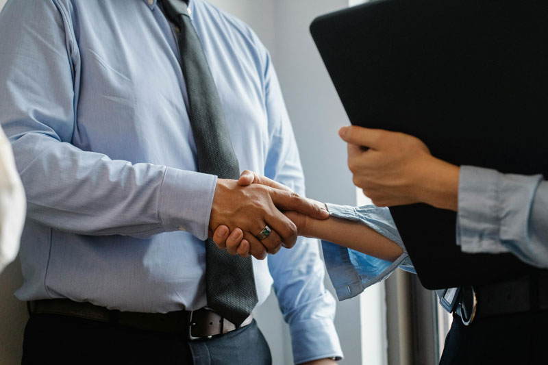 Two professionals shaking hands, with a third person holding a tablet nearby.