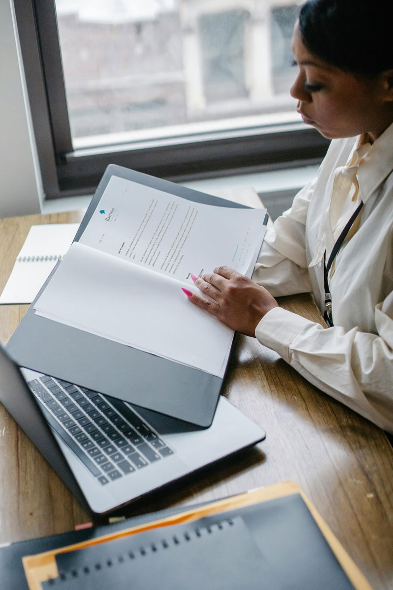 Woman reading a document from a folder, with a laptop open on the desk.