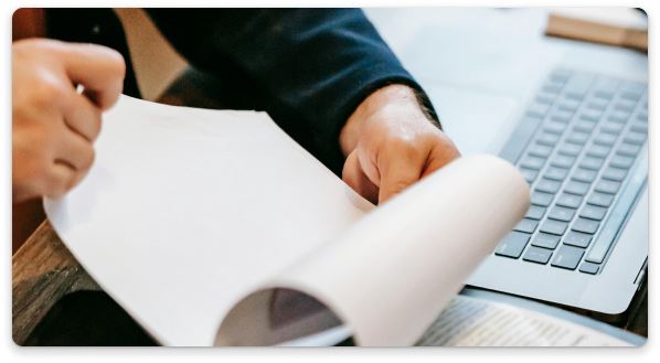 Person flipping through a document next to a laptop on a desk.